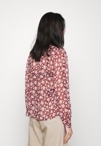 Blusa floreale rossa con tessuto trasparente, maniche lunghe e orlo arrotondato. Presenta un delicato motivo di fiori bianchi su tutta la superficie.