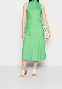 Zielona, bezrękawnikowa sukienka midi z wysokim dekoltem, o gładkiej fakturze i dopasowanym kroju. Połączona z beżowymi klapkami na obcasie i jasną kopertówką.