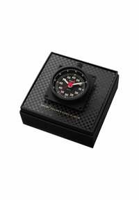 Horloge de bureau noire avec un design carré, aiguilles blanches et rouges, cadran noir et marquages dorés, présentée dans une boîte noire à motif.