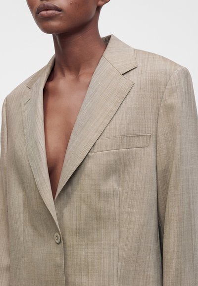 Gros plan d'une personne portant un blazer beige texturé avec un bouton fermé, sans chemise en dessous, sur un fond uni clair.