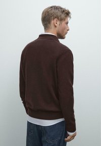 Donkerburgundy gebreide trui met een kraag, geribde manchetten en zoom. Draagt over een wit shirt, gecombineerd met blauwe spijkerbroek.