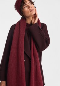 Bordowy zestaw szalika i czapki z prążkowanego materiału, wykonany z miękkiej dzianiny, wyeksponowany z dopasowanym dwurzędowym płaszczem w tym samym kolorze.