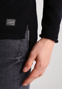 Svart långärmad topp med sidoflik och tygmärke, kombinerad med grå denim, som visar en slät textur och skräddarsydd passform.