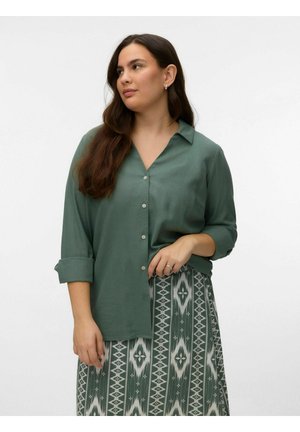 Mujer con cabello largo y castaño viste una camisa verde abotonada y una falda con patrón verde y blanco, de pie frente a un fondo blanco liso.