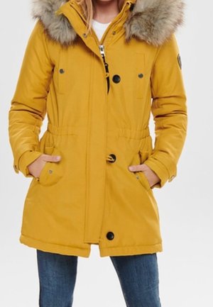 Mujer con abrigo de invierno amarillo mostaza con capucha con borde de piel, cierre de cremallera y botones, con las manos en los bolsillos delanteros, combinado con pantalones vaqueros azules.