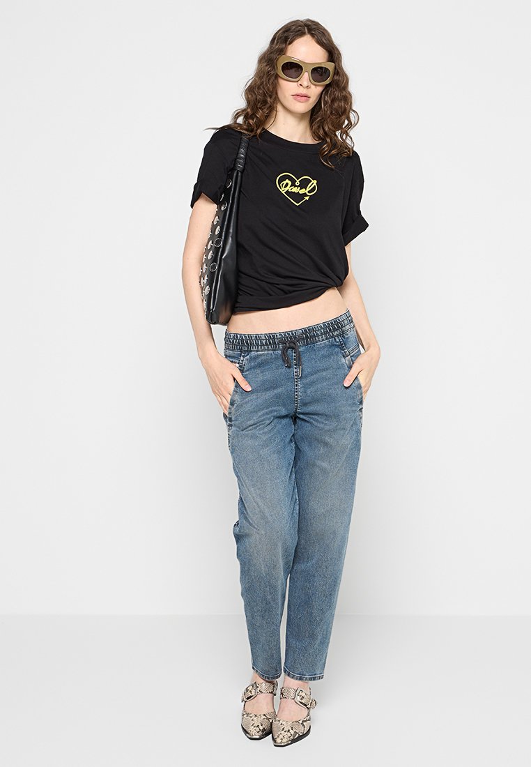 Schwarzes, kurz geschnittenes T-Shirt mit gelbem "Diesel"-Logo, hellblaue Jeanshose mit elastischem Bund, gemusterte beigen Schuhe und schwarze Tasche.