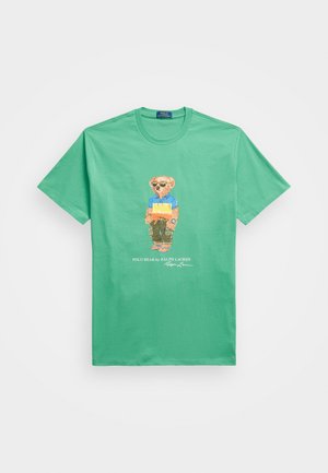 Zielona bawełniana koszulka z kolorową grafiką przedstawiającą niedźwiedzia w koszuli polo i spodniach, z napisem "Polo Bear by Ralph Lauren" poniżej.