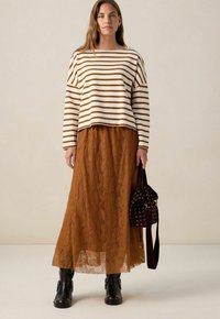 Femme portant un haut à rayures blanches et marron à manches longues, une jupe maxi en dentelle rouille, des bottes noires, et portant un sac en cuir marron foncé avec des œillets.