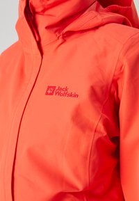 Veste imperméable orange vif avec capuche, présentant une surface texturée et un logo qui indique "Jack Wolfskin" dans une couleur contrastante.