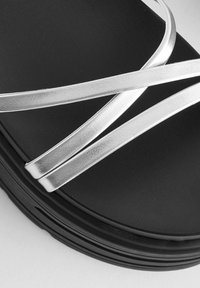 Juodas platforminis šlepetes su metalinėmis sidabrinėmis kryžminėmis juostelėmis. Sklandus paviršius, apvalios formos ir šiek tiek pakilusi padas.