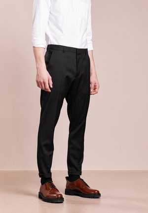Mężczyzna noszący czarne spodnie na miarę, białą koszulę zapinaną na guziki, czarne skarpety i brązowe skórzane eleganckie buty, stojący na beżowej podłodze.