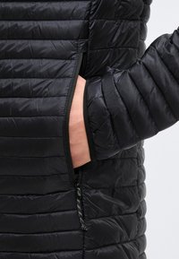 Mână în buzunarul lateral cu fermoar al unei jachete negre matlasate, cu cusături orizontale și un fermoar cu model decorativ.