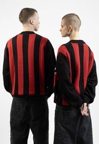 Des pulls en tricot rayés rouges et noirs avec des cols côtelés, associés à un pantalon en denim noir avec des poches texturées. Vue de dos.