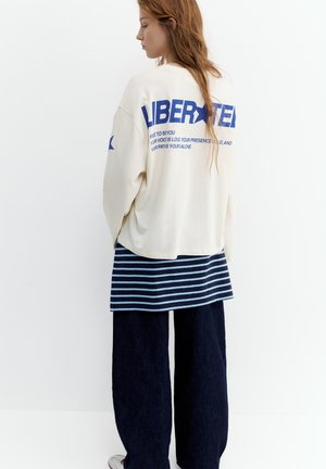 Giovane donna con capelli lunghi indossa un maglione crema oversize con testo blu e stelle, una camicia a righe e pantaloni larghi scuri, in piedi di lato.