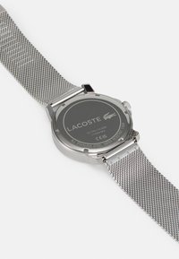 Reloj metálico con una correa de malla de acero inoxidable. Caja redonda de color plateado con la parte trasera negra mate, que presenta texto grabado y un logo. Resistente al agua.