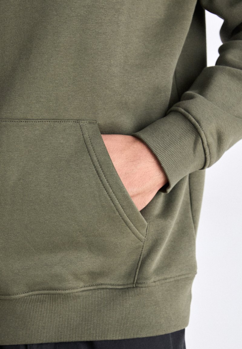 Felpa verde oliva con tasca a marsupio e polsini a costine. Texture morbida e design casual, caratterizzato da una vestibilità rilassata e semplici dettagli delle cuciture.
