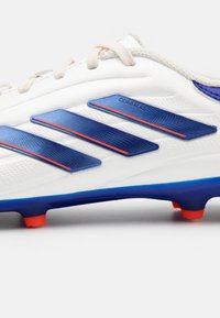 Scarpa da calcio bianca con accenti blu e arancioni, caratterizzata da tre strisce blu e una tomaia sintetica testurizzata per una migliore presa.