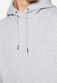 Sudadera gris claro con una superficie texturizada, que cuenta con una capucha con cordón y extremos metálicos en los cordones. Diseño simple y casual.