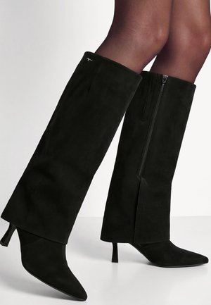Paire de bottes noires en daim montant jusqu'aux genoux, à bouts pointus, zips latéraux, talons de hauteur moyenne, portées avec des collants noirs transparents.