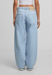 Osoba v světlomodrých širokých džínách a bílém crop topu, zobrazená zezadu na jednobarevném šedém pozadí.