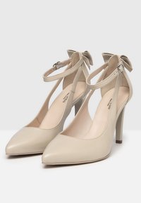 Décolleté beige con punta a punta, cinturino alla caviglia e dettaglio a fiocco; texture in pelle liscia e tacco a stiletto.