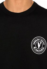 T-shirt in cotone nero con scollo rotondo e un logo circolare bianco con il testo "VERSACE JEANS COUTURE" e una "V" stilizzata al centro.