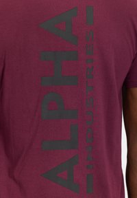 Bordový bavlněný tričko s výrazným černým vertikálním potiskem "ALPHA INDUSTRIES" na zádech. Krátké rukávy, hladká textura.