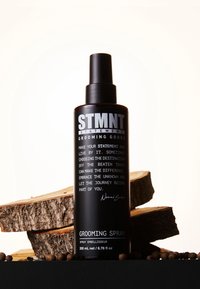 Spruzzatore nero con finitura opaca e forma cilindrica. Presenta testo e logo bianchi. Posato su ceppi di legno con sfondo scuro.