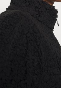 Zwarte fleece jas met een gestructureerd, fluffy oppervlak. Voorzien van een hoge kraag en een ritssluiting voor warmte en comfort.
