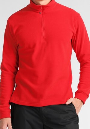 Roter Fleecepullover mit langen Ärmeln und Viertel-Reißverschluss am Kragen, getragen mit schwarzer Hose, Hand in der Tasche ruhend.