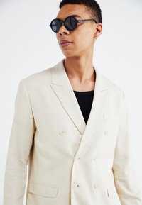 Blazer de lino beige de doble botonadura con solapas con muesca, dos botones y un bolsillo en el pecho. Lleva puesto un top negro con superficie acanalada y gafas de sol redondas.