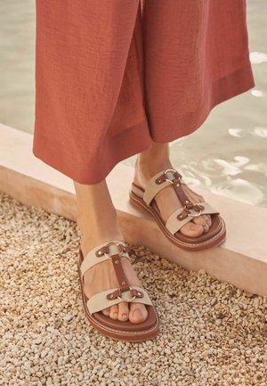 Picioare purtând sandale din piele bej și maro cu inele metalice, stând pe pietriș lângă marginea apei, pantaloni de culoare ruginie tăiați deasupra gleznelor.