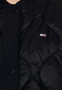 Black quilted jacket with rounded collar, featuring textured sections, button closure, and a small logo in red, white, and blue.