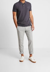 Polo shirt gris foncé avec trois boutons et un petit logo, associé à un pantalon gris clair retroussé aux chevilles et des chaussures blanches avec des semelles marron.