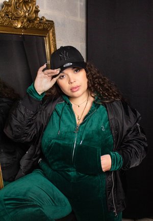 Groene fluwelen jumpsuit met voorzakken, gelaagd onder een zwarte bomberjack. Model draagt een zwarte baseballpet en gouden kettingen.