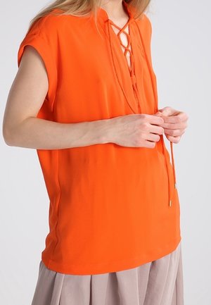 Femme ajustant l'encolure lacée d'un top sans manches orange vif associé à une jupe beige sur un fond uni.