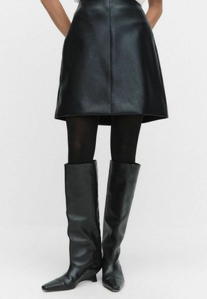 Mini-jupe en cuir noir avec une texture lisse et un design en A, associée à des bottes en cuir noir hautes avec des bouts pointus et une coupe ajustée.