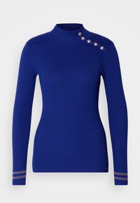 Ribbstickad blå tröja med hög krage, långa ärmar och guldfärgade knappar på axeln, med metalliska randiga detaljer på muddar.