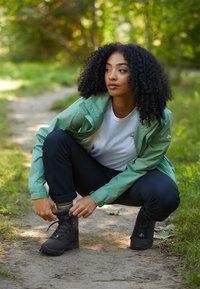 Kvinna med lockigt hår som sitter på huk på en grusväg utomhus, justerar sin svarta vandringskänga, bär en grön jacka, vit skjorta och svarta byxor.