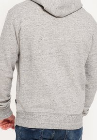 Szara bluza z kapturem wykonana z teksturowanego materiału typu fleece, z regulowanym kapturem i ściągaczem na dole. Posiada długie rękawy i małe logo przy dolnej krawędzi.