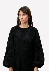Blouse noire en tissu transparent avec un motif floral en relief, dotée d'une encolure arrondie et de manches bouffantes volumineuses.