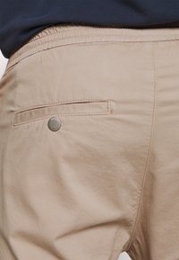 Lichtbeige stoffen broek met een elastische tailleband, zijzakken en een achterzak met een metalen knopf detail. Zachte textuur.