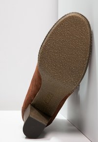 Brązowy zamszowy but z teksturowaną gumową podeszwą, wyróżniający się piętą w stylu stacked oraz detalem przeszycia po bokach. Spód jest lekko zużyty.