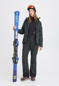 Skidress med svart jakke, svart termisk topp og mørke skibukser. Modellen holder blå ski med bindinger og har på seg en svart lue og goggles.