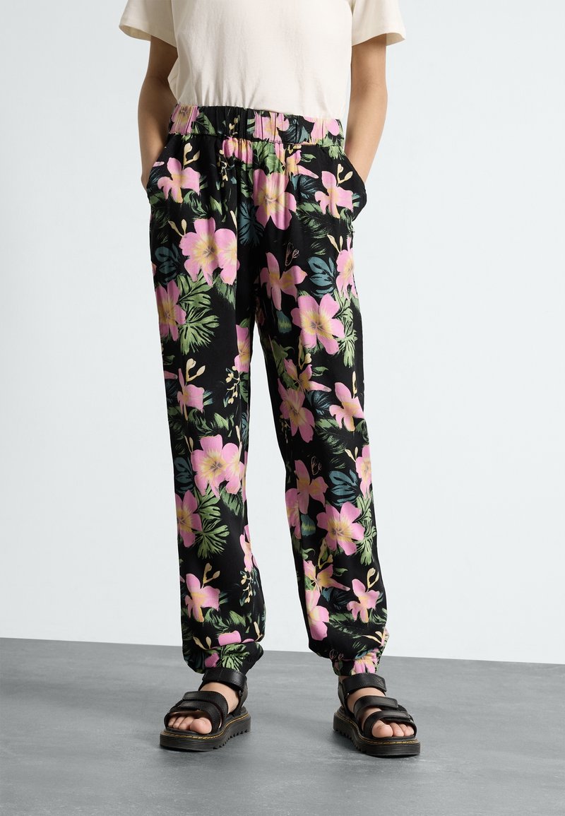 Personne portant un pantalon noir avec un motif floral rose et vert, taille et poignets élastiques, assorti à des sandales noires et un haut beige.