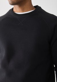 Man in een zwart sweatshirt over een wit T-shirt, met een ronde hals en gestikt detail bij de kraag.
