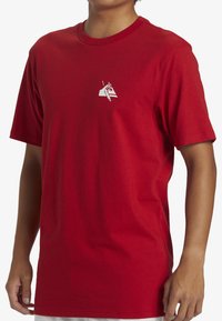 Camiseta de algodón roja con cuello redondo y mangas cortas. Presenta un pequeño gráfico blanco en el lado izquierdo del pecho. Corte recto con una textura suave.