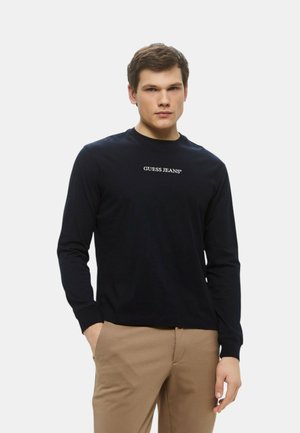 T-shirt a maniche lunghe in cotone nero con collo rotondo, presenta il logo "GUESS JEANS" in bianco centrato sul petto. Texture morbida, design semplice.