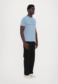 Gaiši zils īsās piedurknes T-krekls ar melnu "A|X ARMANI EXCHANGE" logotipu, apvienots ar melnām biksēm un bēšiem sporta apaviem. Ikdienišķa piegriezne.