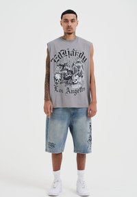 Camiseta gris sin mangas con estampado gráfico, combinada con shorts de denim azul claro con bordado lateral. Zapatillas blancas completan el look.